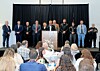 Officials stand on a stage behind a podium as people sit at tables in front of them.