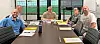 Four men seated around a conference table in a conference room with laptops, papers, and name placards.
