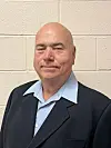 Bald man wearing a dark blazer and light blue shirt stands against a beige cinder block wall.