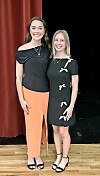 Two women posing side by side on a stage in front of red curtains.