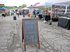 Chalkboard welcome sign at an outdoor farmers market with vendor tents and tables.