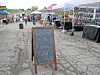 Chalkboard welcome sign at an outdoor farmers market with vendor tents and tables.