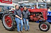 Two workers stand beside a vintage red tractor with large rear wheels in a workshop.