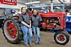 Two workers stand beside a vintage red tractor with large rear wheels in a workshop.