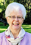 Smiling elderly woman with white hair and glasses standing outdoors in a park.