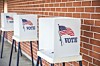 Voting booths with American flags and the word VOTE lined up beside a brick wall.