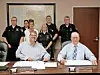 Group of officials standing behind two seated men at a conference table in a conference room.