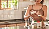Woman adding powder to a shaker bottle on a kitchen counter with jars and a glass nearby.