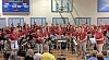 A large marching band in red shirts performs in a gymnasium with drums, brass, and woodwinds.