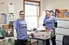 Two people in matching shirts stand next to stacked boxes in an office filled with supplies.