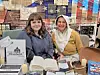 Two women seated behind a table displaying handmade items, an open book, candles, and brochures.