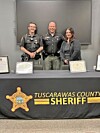 Two sheriff's deputies and a woman stand behind a Tuscarawas County Sheriff table covered with a black banner and certificates.