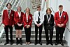 Six students stand posing on a staircase inside a school building, wearing red blazers and formal uniforms.