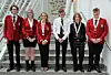 Six students stand posing on a staircase inside a school building, wearing red blazers and formal uniforms.