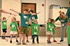 Children with green lanyards stand with arms outstretched during a group activity indoors.