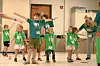 Children with green lanyards stand with arms outstretched during a group activity indoors.