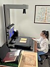 Woman seated at a large-format scanner in a room with maps and document boxes.