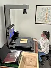Woman seated at a large-format scanner in a room with maps and document boxes.