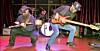 Two musicians play electric guitars on a stage with drums and curtains behind them.