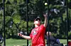 Tennis player in red shirt reaching up with racket to hit a ball on an outdoor court.