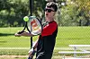 A tennis player swings a racket on an outdoor court beside a chain-link fence and bleachers.