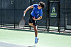 Tennis player in blue mid-serve on an outdoor court near a fence with a logo.