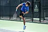 Tennis player in blue mid-serve on an outdoor court near a fence with a logo.