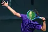 Tennis player in purple shirt raising one arm and swinging a racket during a match.