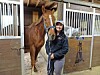 Woman holding a horse’s lead rope in a stable beside a brown horse with a white blaze.