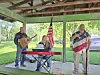 Three people perform under a pavilion with keyboard, guitar, and microphone near an American flag.