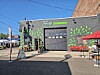 A storefront with green leaf artwork on the wall, a gray door, and tables with umbrellas outside.