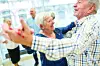 Older adults dancing together indoors in a bright room, with one man in the foreground and a smiling woman beside him.