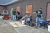 Workers stand beside a brick building, fitting and finishing large wall panels on a concrete walkway.