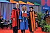 Three people in academic regalia pose on a commencement stage with seating and plants in the background.