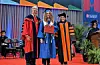 Three people in academic regalia pose on a commencement stage with seating and plants in the background.
