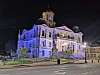 Historic building illuminated in blue and white at night with a monument and street in front.