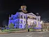 Historic building illuminated in blue and white at night with a monument and street in front.