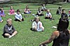 Children sitting on grass in a park, with an adult facing them in the foreground.
