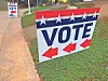 Large yard sign reading VOTE with stars and red arrows beside a sidewalk.