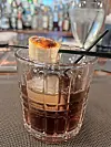 A short glass of dark cocktail with a toasted marshmallow garnish and black straw on a bar counter.