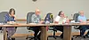 Several people seated at a long table in a meeting room with laptops and papers.