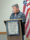 A man speaks at a podium beside an American flag in an indoor room.