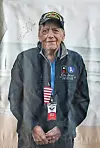 An older man in a veteran cap and dark jacket stands in front of a light backdrop.