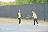 Two tennis players exchange shots on an outdoor court near a dark fence.