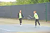 Two tennis players exchange shots on an outdoor court near a dark fence.
