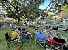 People seated in folding chairs on a grassy park lawn under trees at an outdoor gathering.
