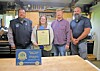 Four people standing in a workshop, one holding a framed certificate beside an award plaque on a table.