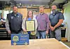 Four people standing in a workshop, one holding a framed certificate beside an award plaque on a table.
