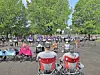 People seated in folding chairs and standing on a basketball court in a park with trees in the background.