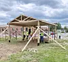 Workers build a wooden pavilion frame on a grassy lot beside a brick building.